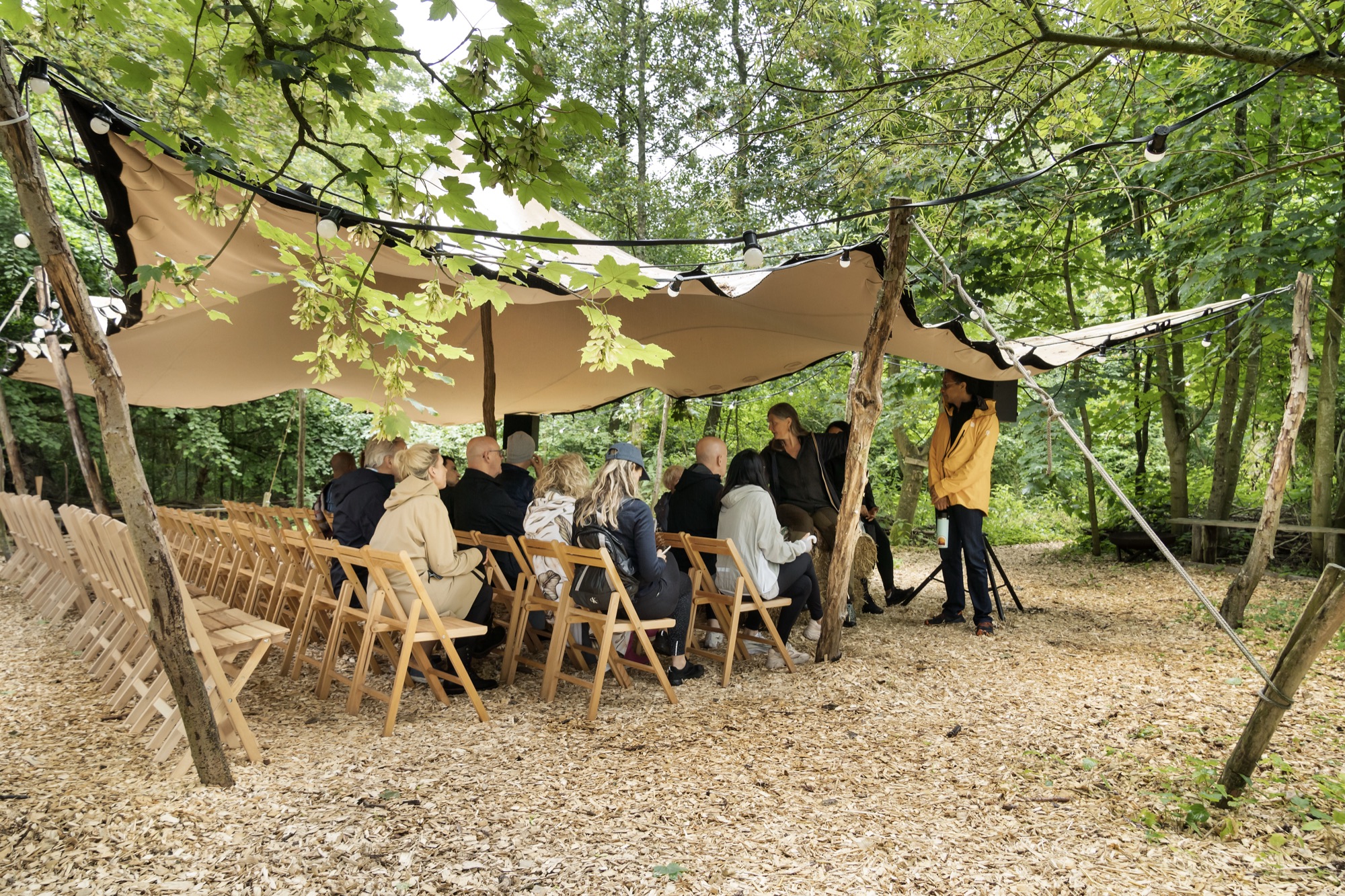 A group in conversation under a canvas roof among the trees